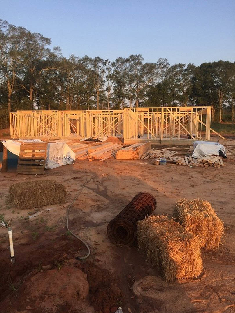 Wood-framed house under construction with stacked hay bales on dirt ground, surrounded by trees and plants under a clear sky