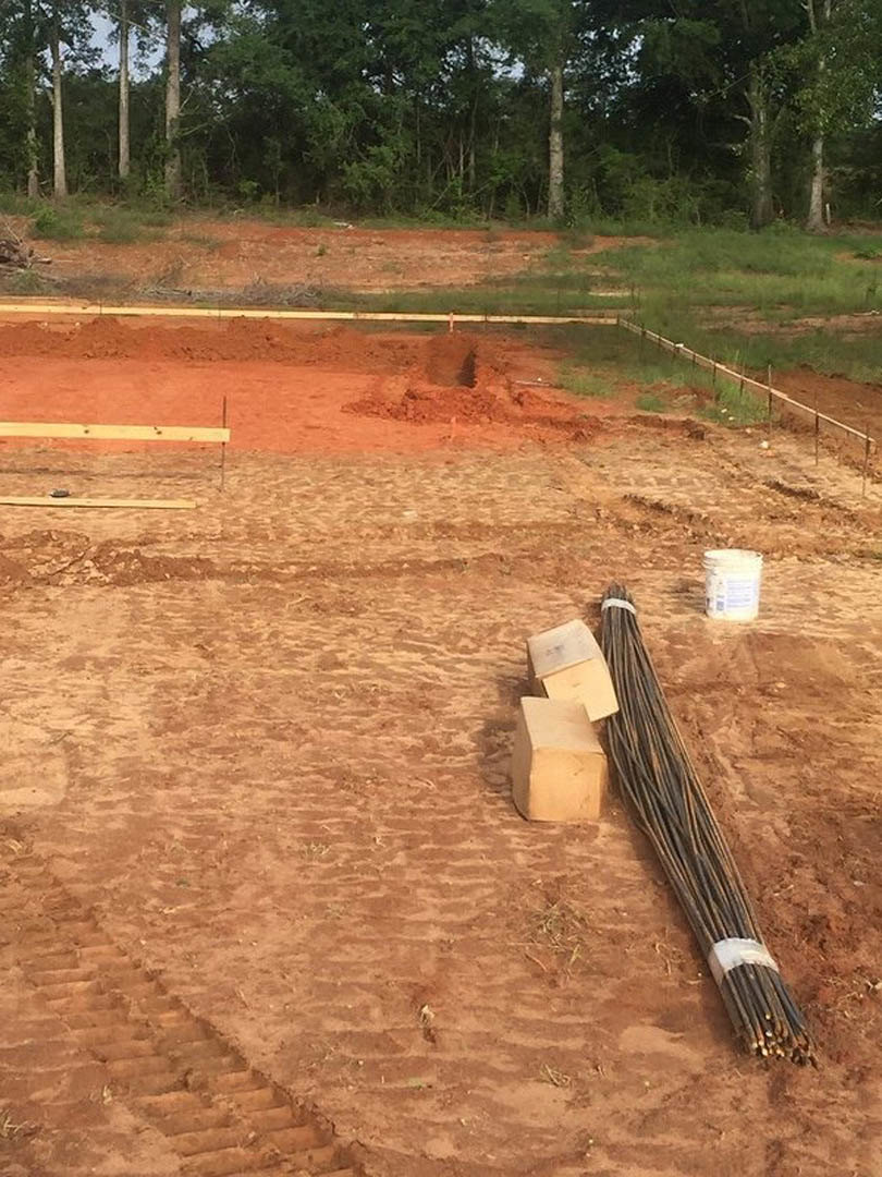 Construction site with scattered wood planks, coiled black wire, white bucket labeled in blue, and dirt ground bordered by a metal fence and trees.