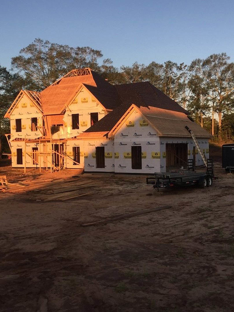 Framed house under construction with exposed wood, dirt lot, utility trailer parked in front, trees and blue sky in background
