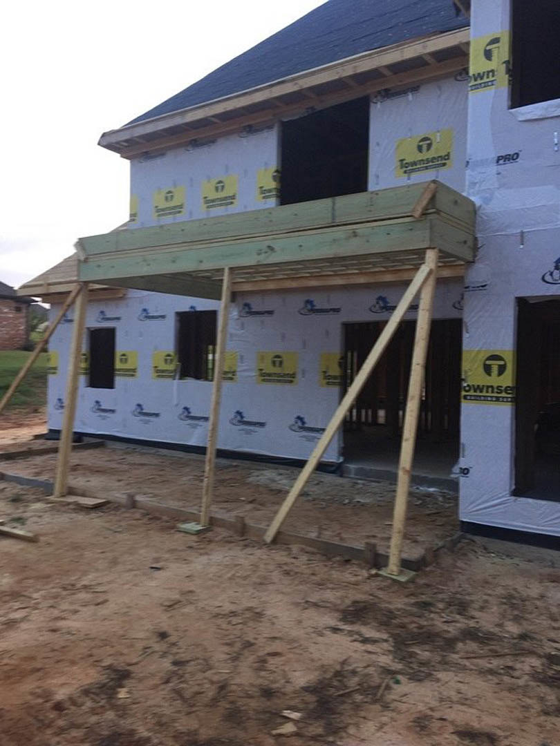 Wood-framed house under construction with exposed beams, dirt ground, yellow caution sign, and ladder against exterior structure.