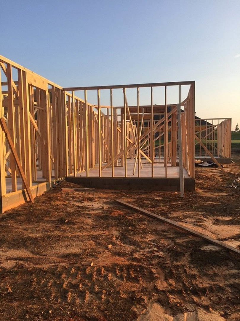 Wooden framing structure of a house under construction with exposed beams and plywood, set on a dirt lot under a clear sky.