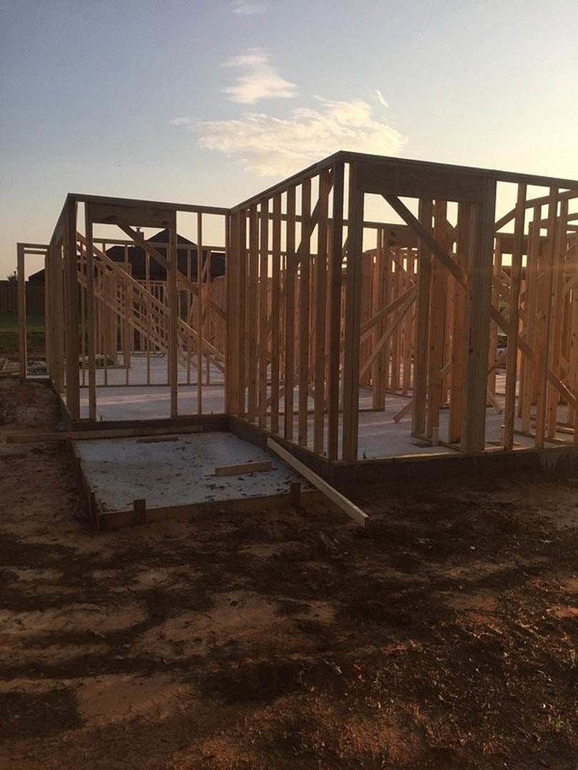 Wooden framing and concrete foundation of a house under construction, exposed dirt floor, partial staircase, blue sky with scattered clouds in background.