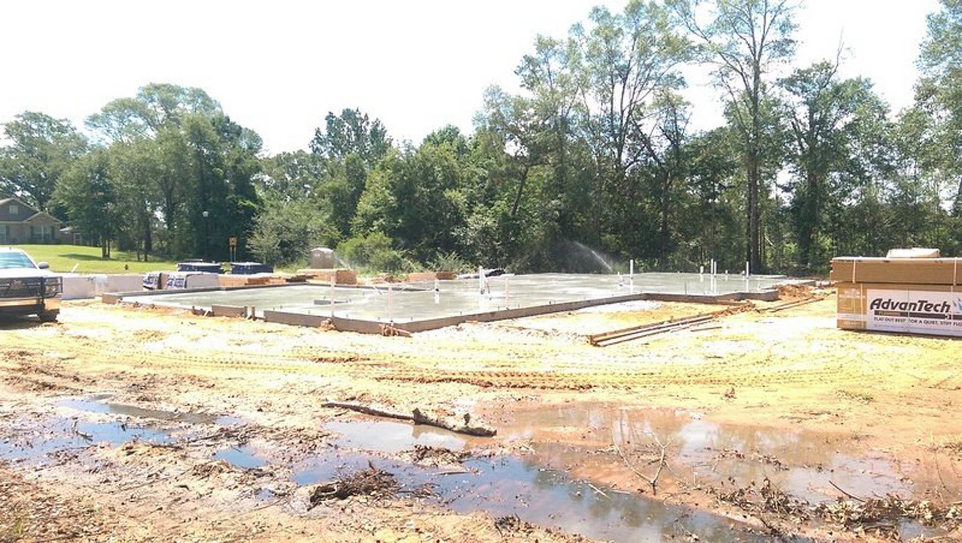 Muddy construction site with concrete foundation walls, standing water, grassy patches, and scattered building materials
