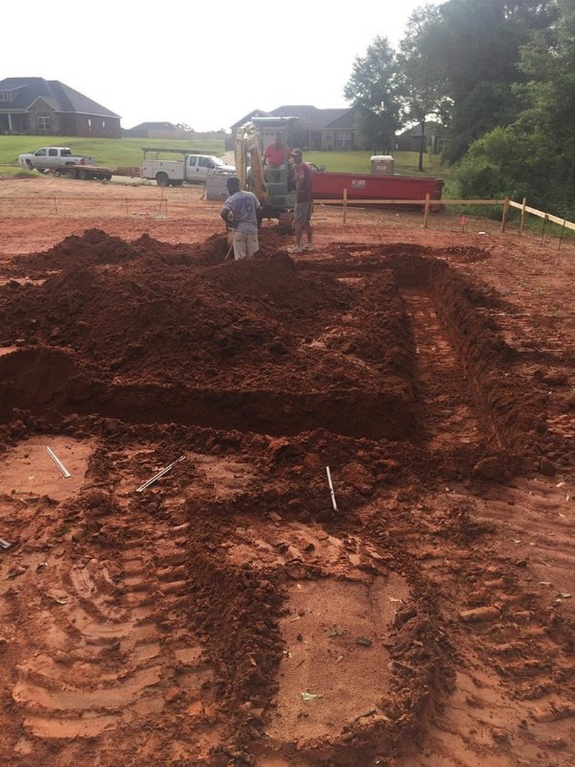 Men in work clothes standing on dirt near a partially built house with exposed roof, white truck parked beside green field, trees and blue sky in background