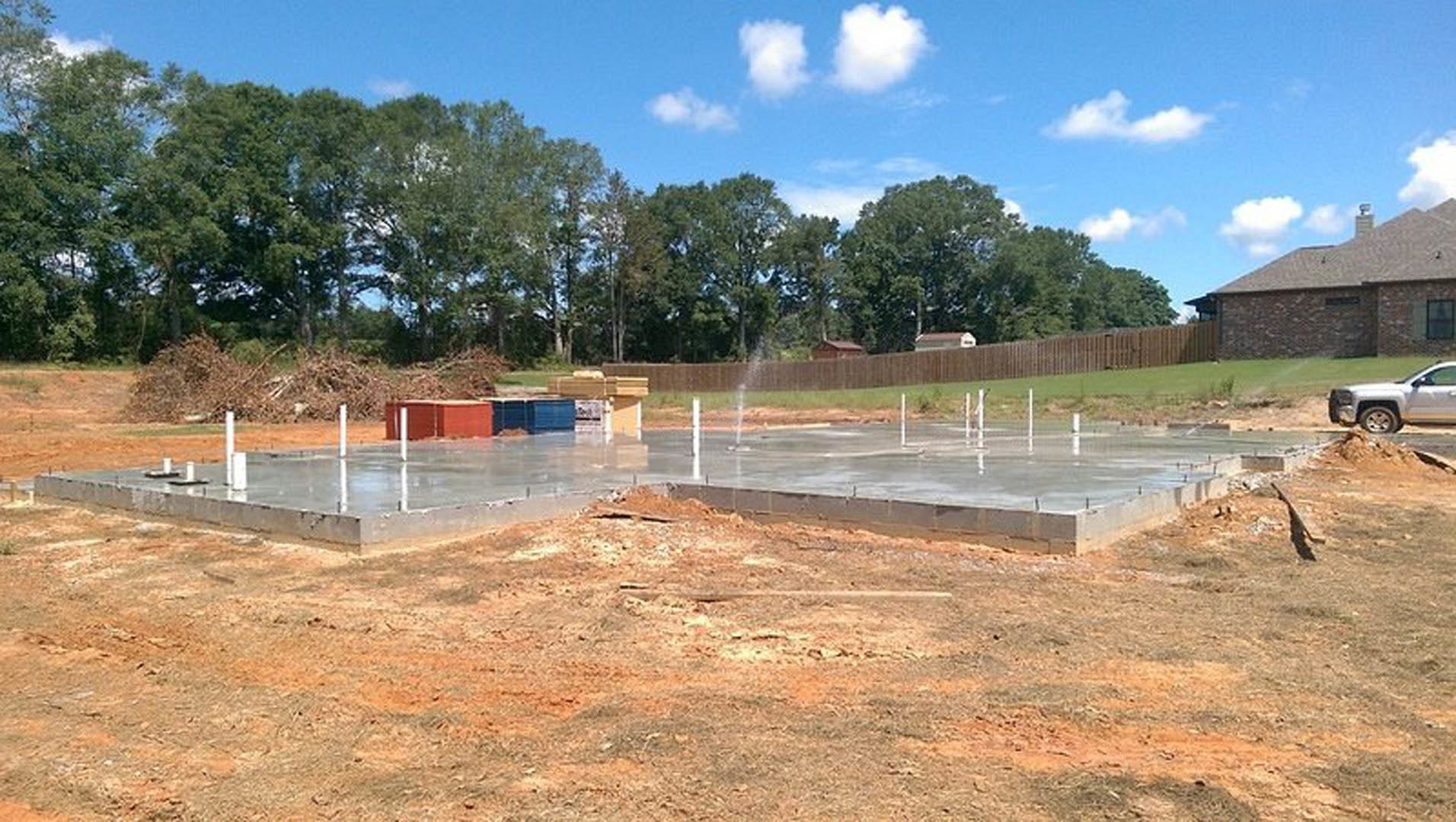 Dirt lot with construction materials, wooden fence, and mature trees in background; partially built house with chimney visible