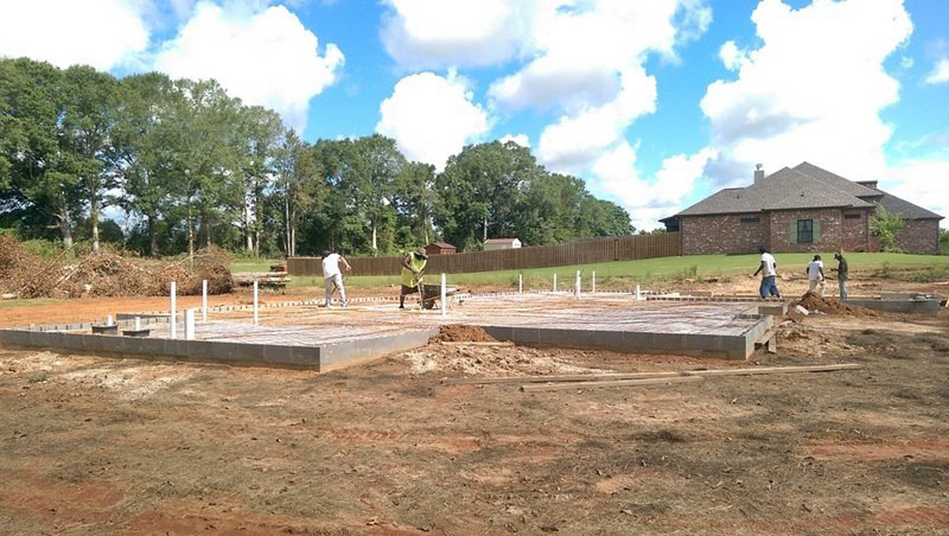 Framed custom home under construction with exposed wooden beams, workers on dirt lot, scattered building materials, cloudy sky, and trees in background