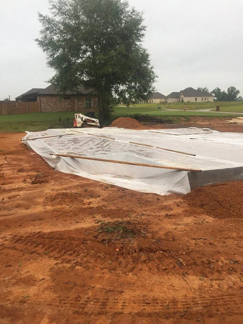 White tarp draped over construction site, brown brick wall with black roof, tree and grass in foreground, white and red vehicle parked nearby