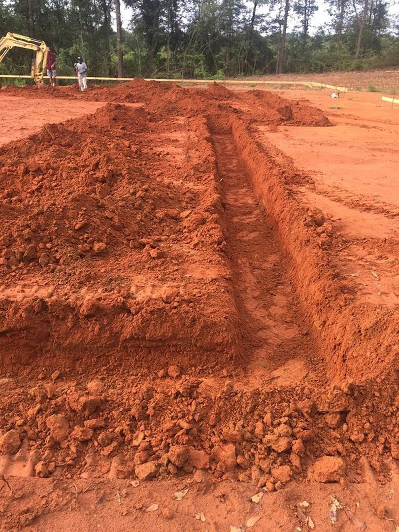 Wide trench dug into red-brown soil with a yellow construction vehicle nearby, surrounded by dense forest trees and muddy ground.