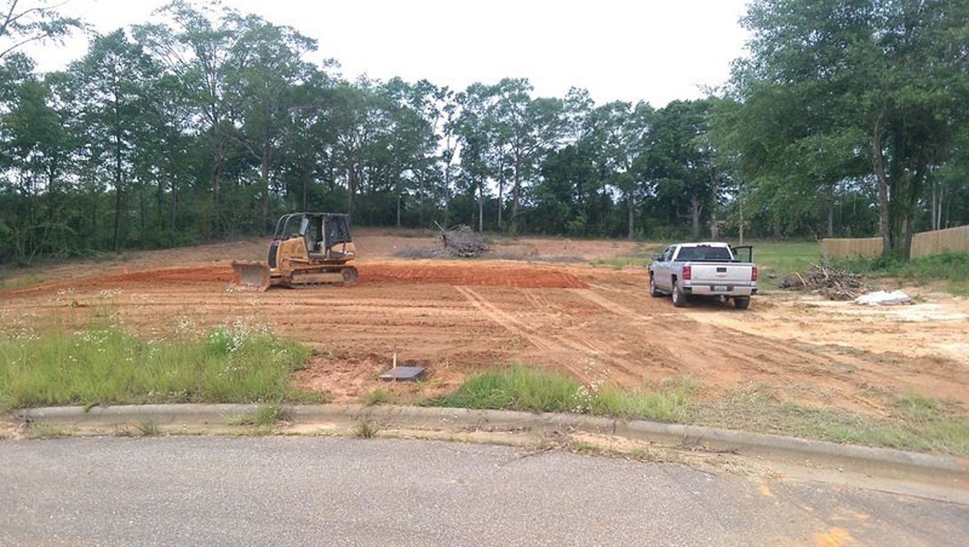 White pickup truck and yellow bulldozer parked on dirt construction site bordered by grassy field and trees under clear sky