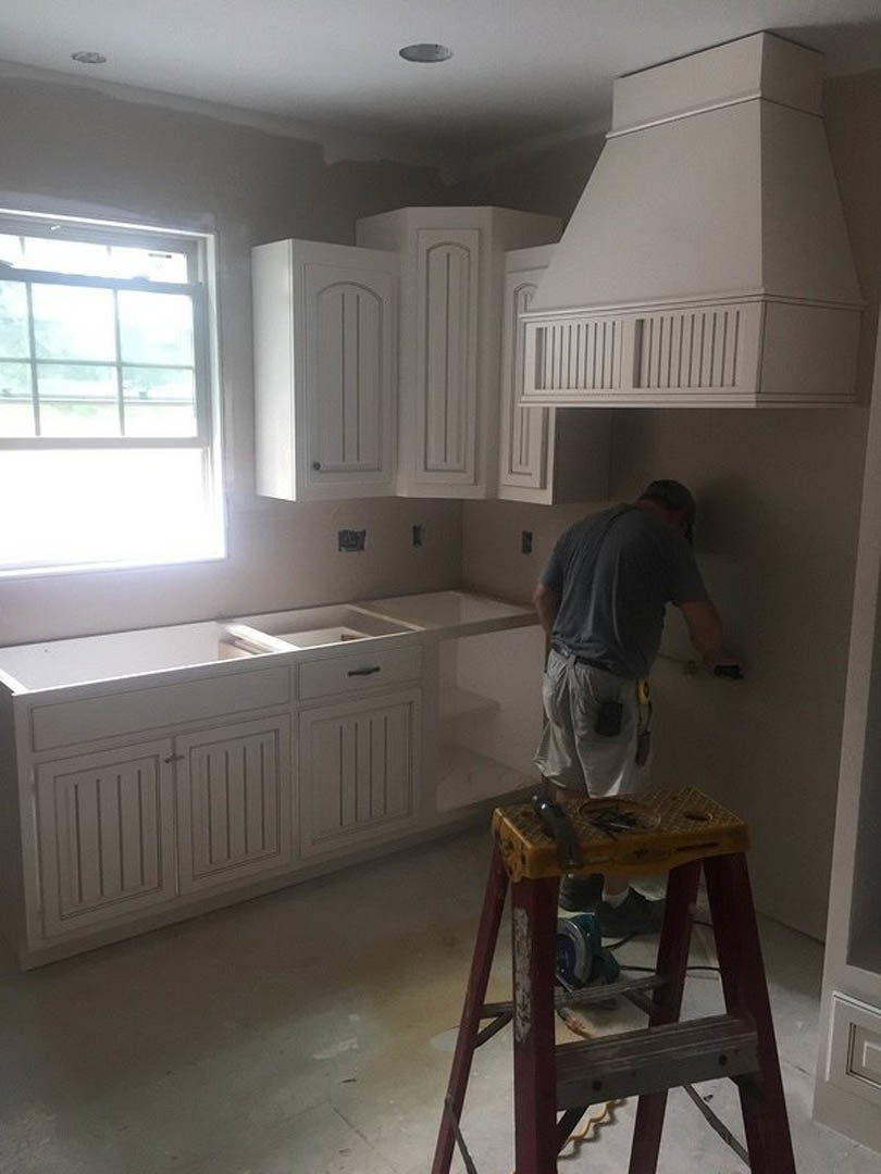 Man in grey shirt painting kitchen wall beside white cabinets with open top, stepladder on light floor, multi-pane window in background, phone visible in pocket.