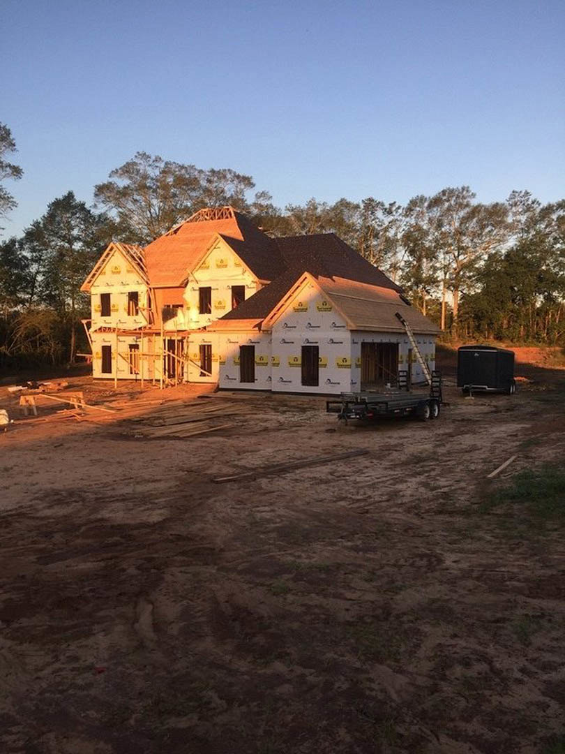 Framed house under construction with exposed wooden beams, partially installed roof, dirt lot, metal trailer with black box nearby, clear blue sky and scattered trees in background