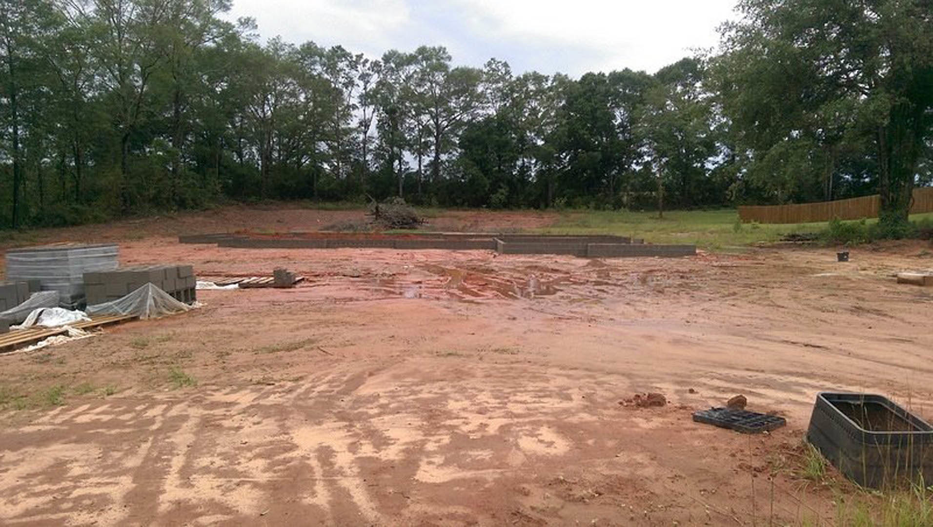 Dirt lot bordered by mature trees, brick wall along one side, rectangular object on grass, cloudy sky overhead
