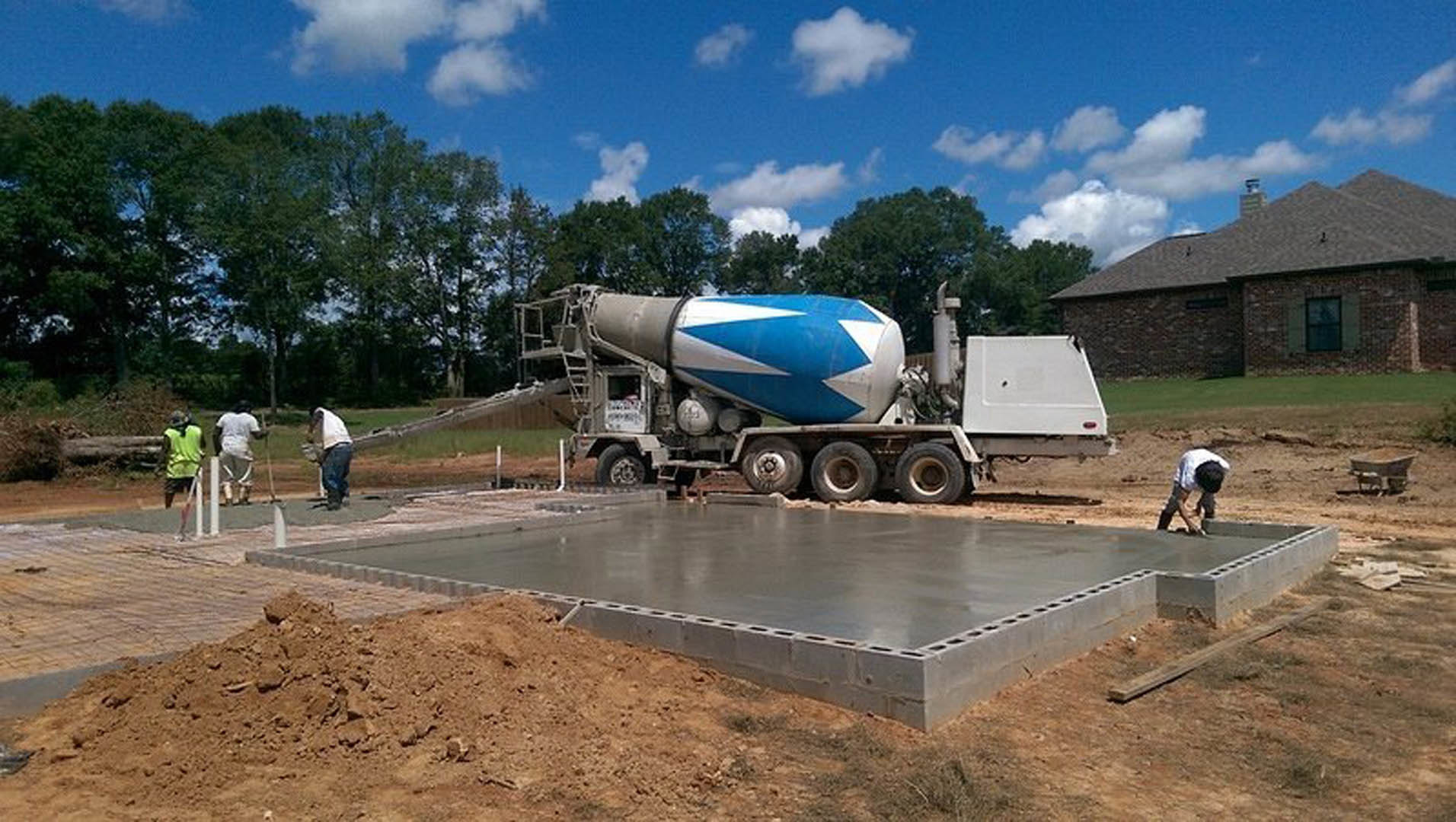 Man in white shirt and blue jeans standing beside large concrete mixer truck on gravel driveway, trees and cloudy sky in background, construction materials scattered nearby
