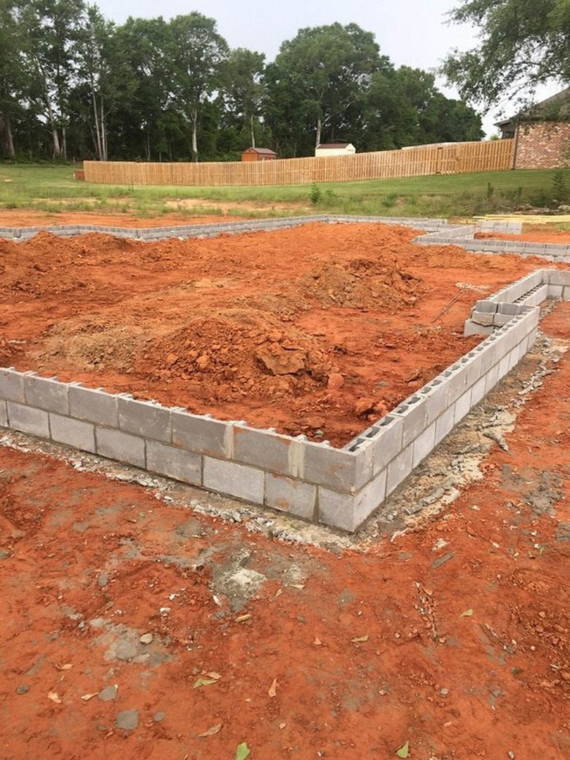 House foundation under construction with exposed dirt and stacked bricks, surrounded by trees, a fence, and a nearby building