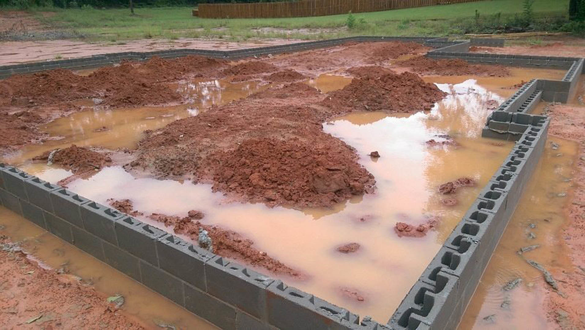 Wet concrete foundation with muddy puddles, surrounded by soil, grass, and scattered construction debris
