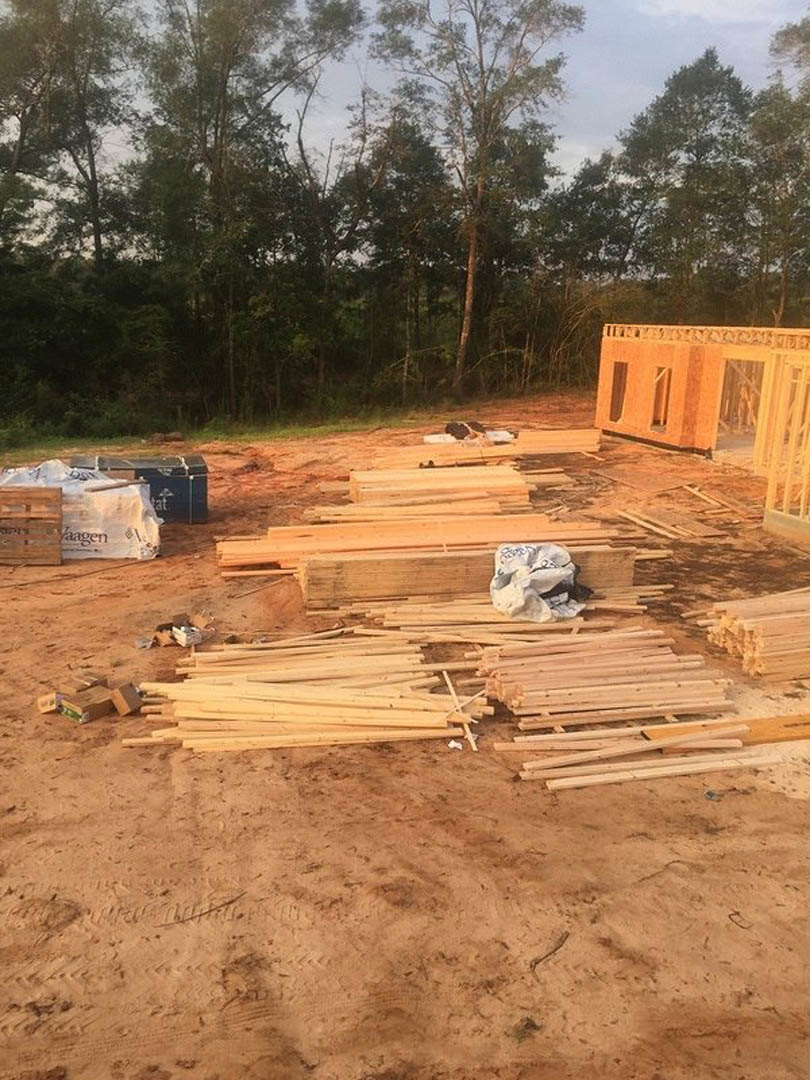 Wood-framed house under construction with exposed lumber, foundation, piles of wood on soil, white tarp with graffiti, and surrounding trees