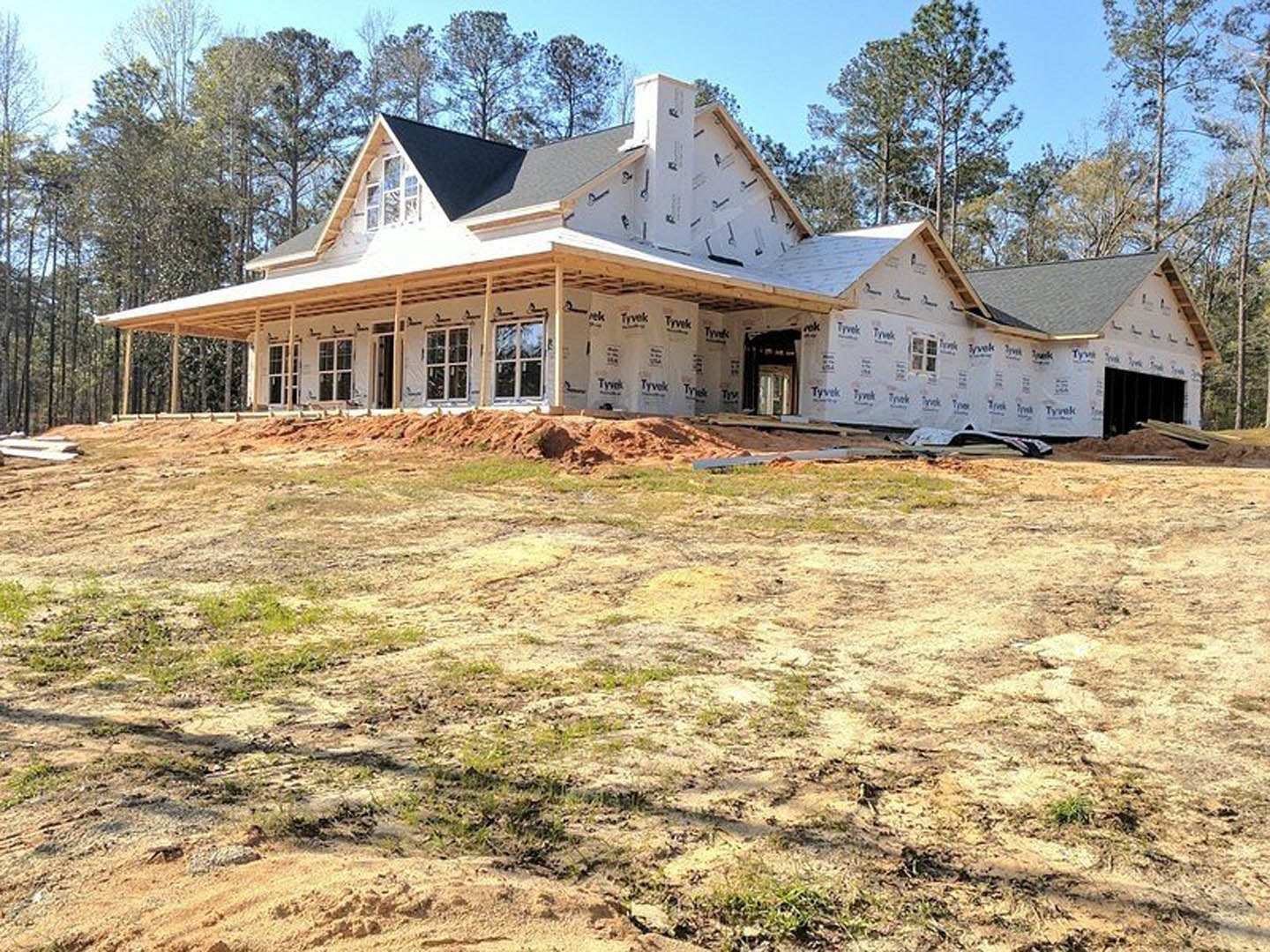 Wood-framed house under construction with spacious covered porch, exposed beams, brick chimney, dirt lot in foreground, and unfinished windows.