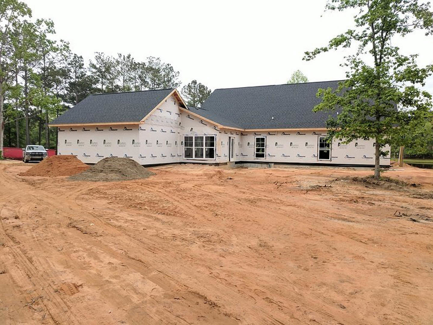 Two-story house under construction with exposed roof, dirt lot in foreground, white truck parked near unfinished exterior, large tree partially obscuring front, visible window