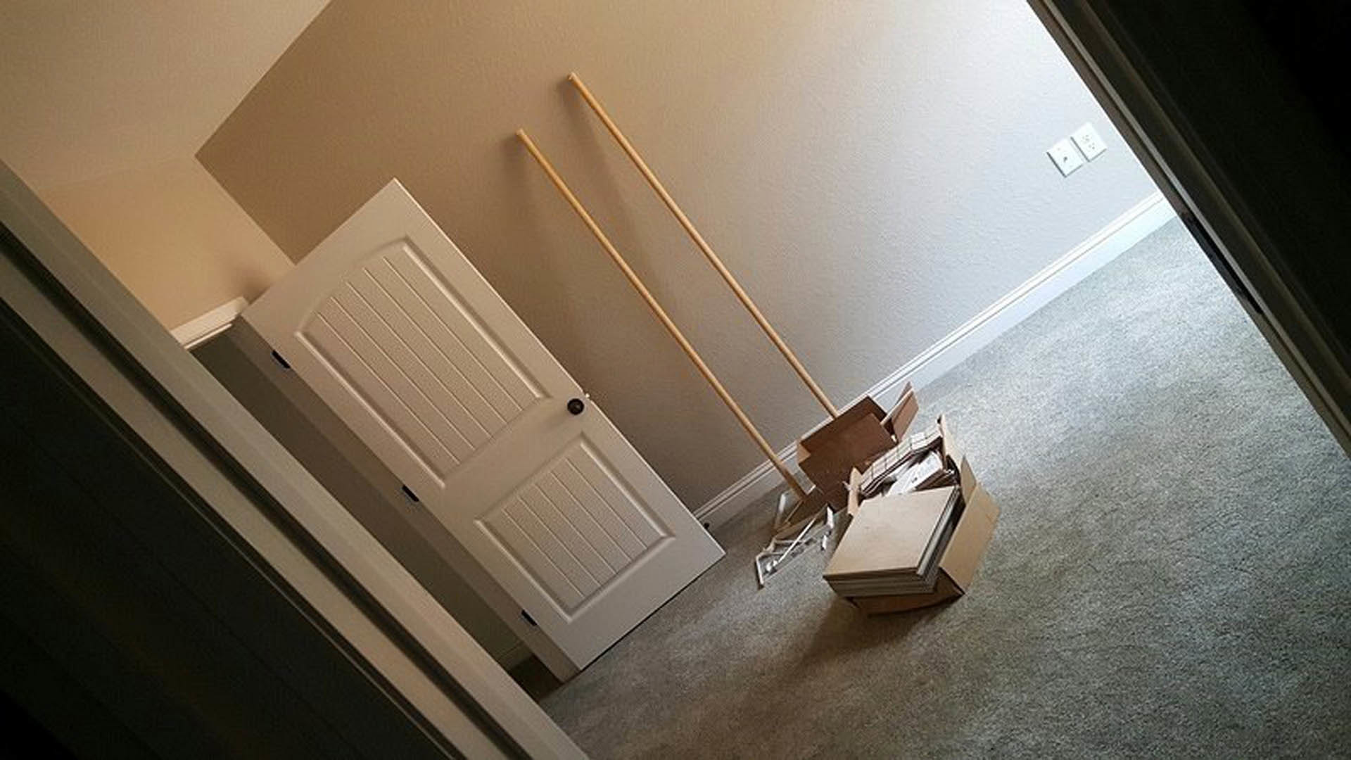 White interior door beside a staircase, stack of pizza boxes on the floor, broom leaning against wall, white electrical outlet visible.