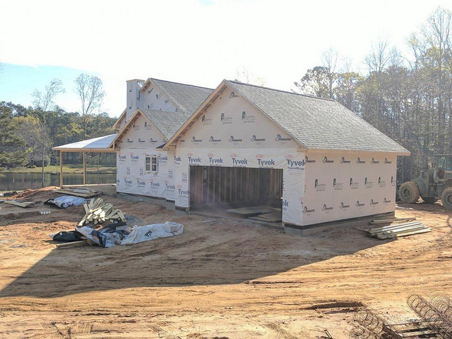 Wood-framed house under construction with attached garage, exposed plywood walls, roof trusses, and construction equipment on dirt lot