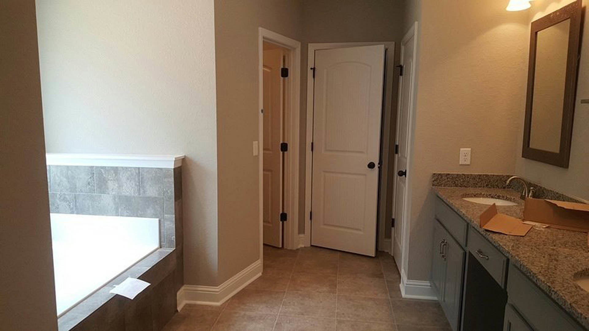 Bathroom with light gray tile floor, white vanity and sink, black faucet, two white doors with black handles, wall-mounted light fixture, white electrical outlet on wall