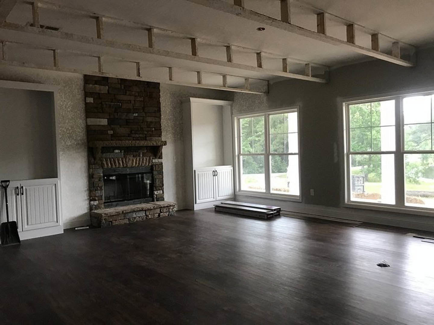 Dark wood flooring and white cabinetry with black handles in a den featuring a central fireplace, large windows framing leafy trees, and metal accents on furniture.