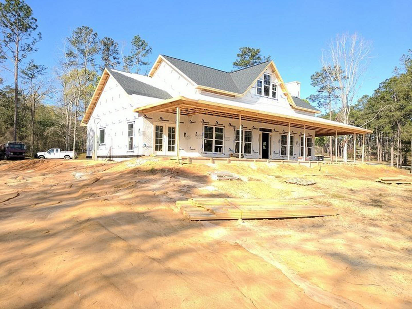 Partially built house with exposed wooden framing and roof, surrounded by tall trees, construction materials and a pickup truck parked nearby