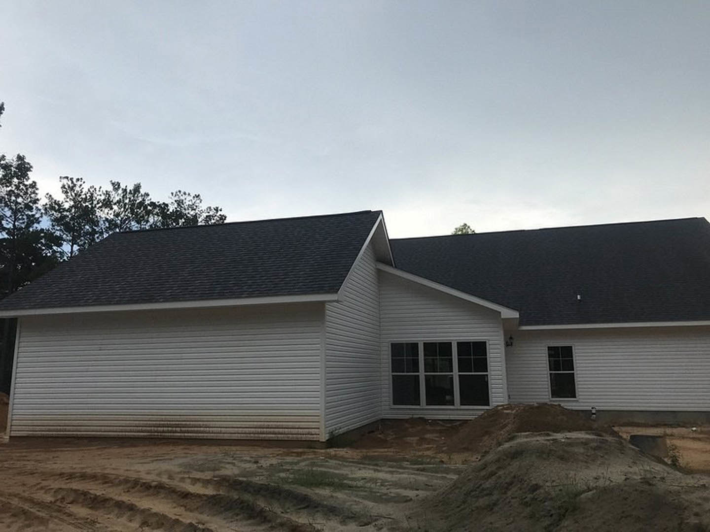 Partially built house with black roof, white-framed windows, light siding, surrounded by dirt patch and mature trees under cloudy sky