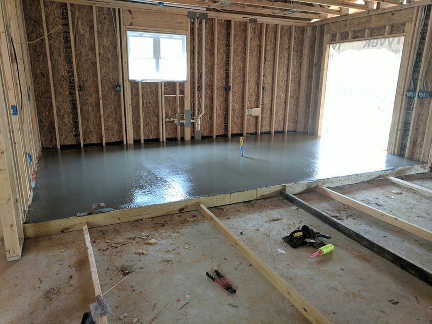 Unfinished room with exposed concrete floor, scattered construction tools, black hat, window letting in natural light, visible insulation and lumber along walls