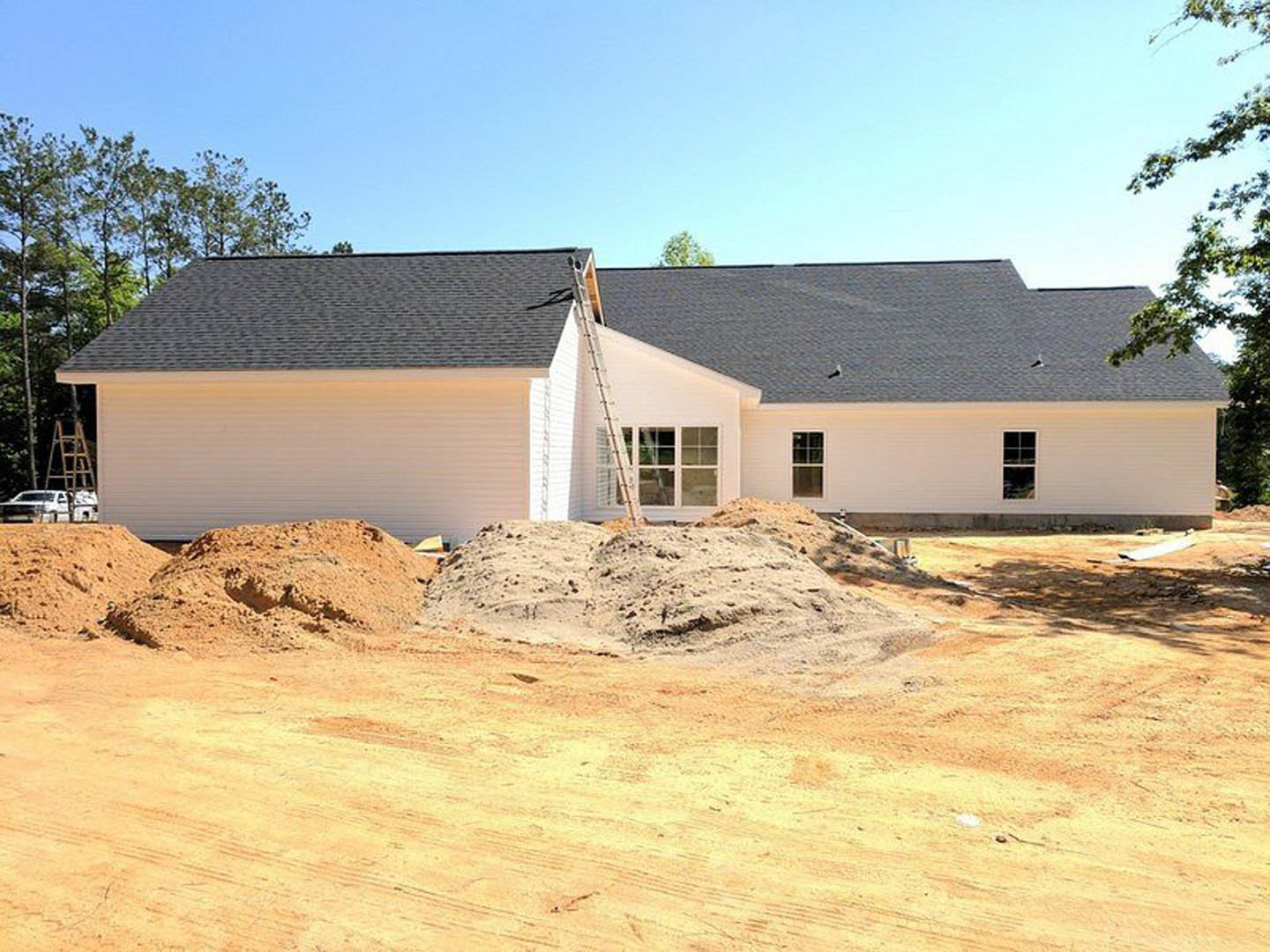 Wood-framed house under construction with exposed sheathing, a ladder leaning against the exterior wall, and a large pile of dirt in the foreground under a clear blue sky
