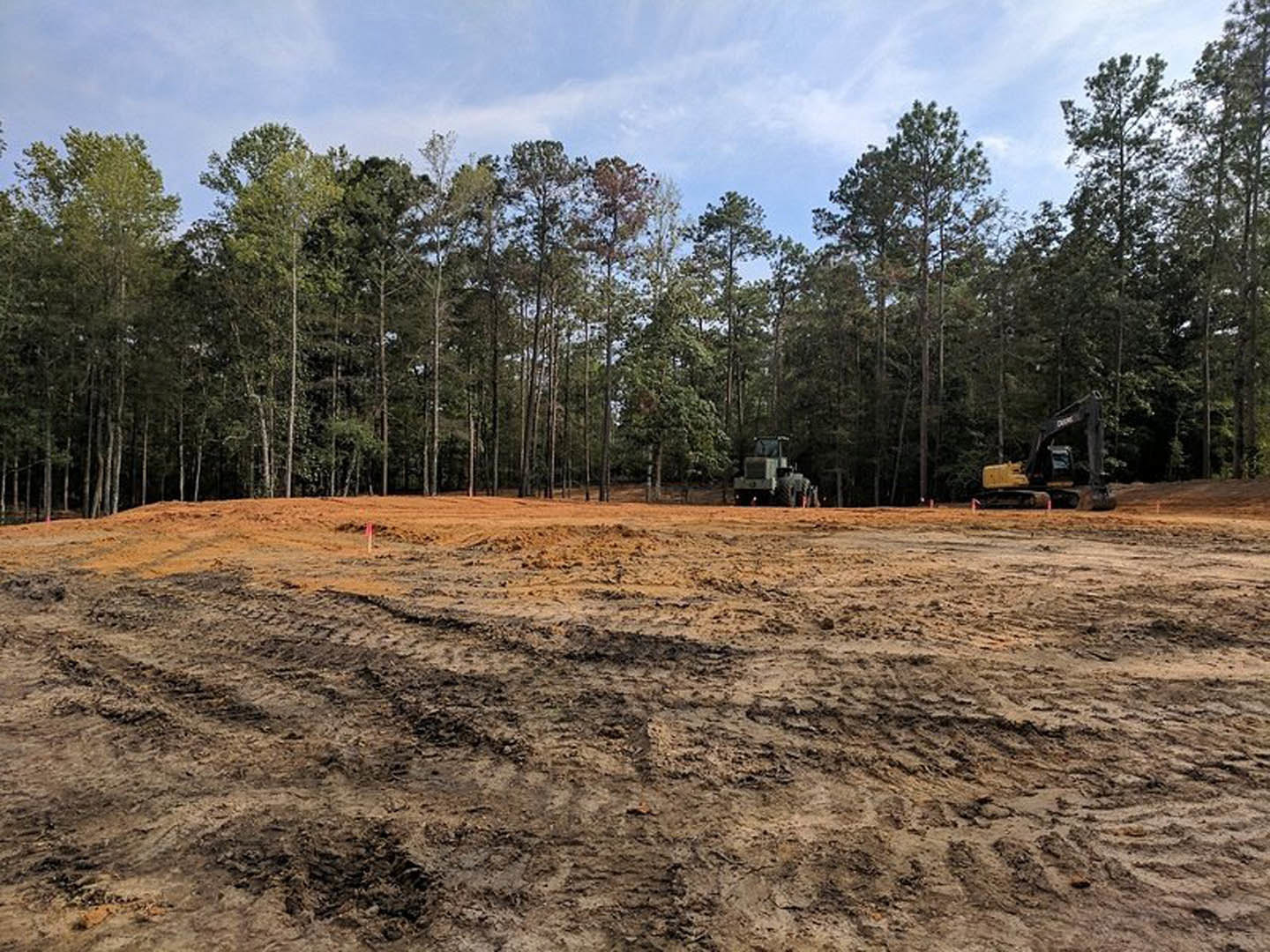 Dirt field with tire tracks, red and white pole, tractor with large wheels near trees under cloudy sky