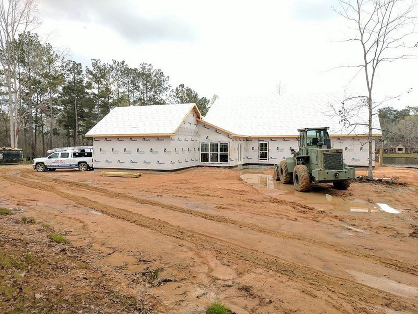 Green tractor parked on muddy dirt road in front of partially constructed house with exposed framing, white truck with ladders nearby, leafless tree, and unfinished pool.