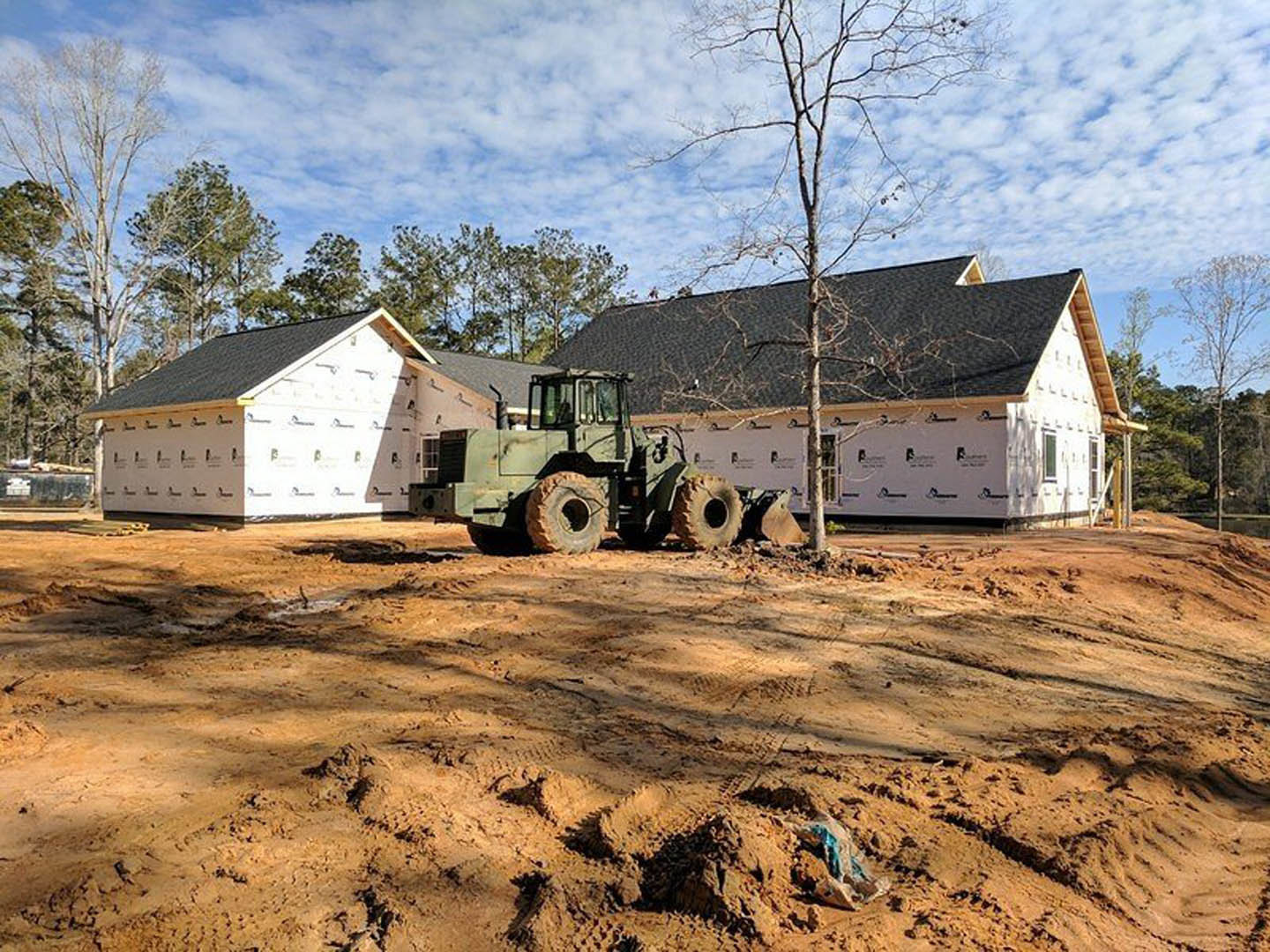 Green tractor parked on dirt driveway in front of modern house with white siding, leafless tree nearby, blue sky and scattered clouds overhead