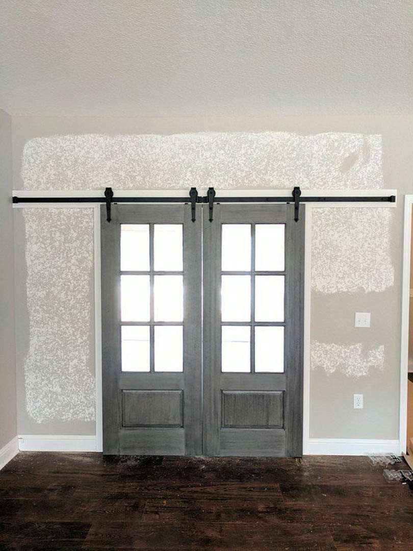 Double entry doors with glass panes beside a rustic sliding barn door, wood flooring, white plaster walls, and a light switch.