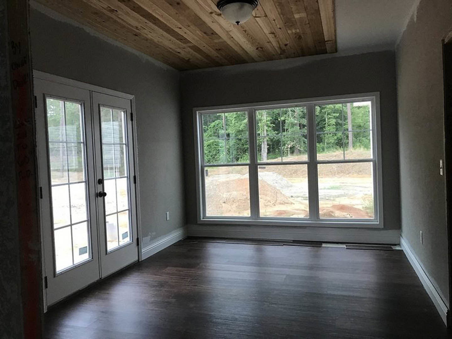 Wood plank flooring, white walls, large windows with tree views, double glass-paneled doors, wooden ceiling with recessed light fixture, modern lamp in foreground
