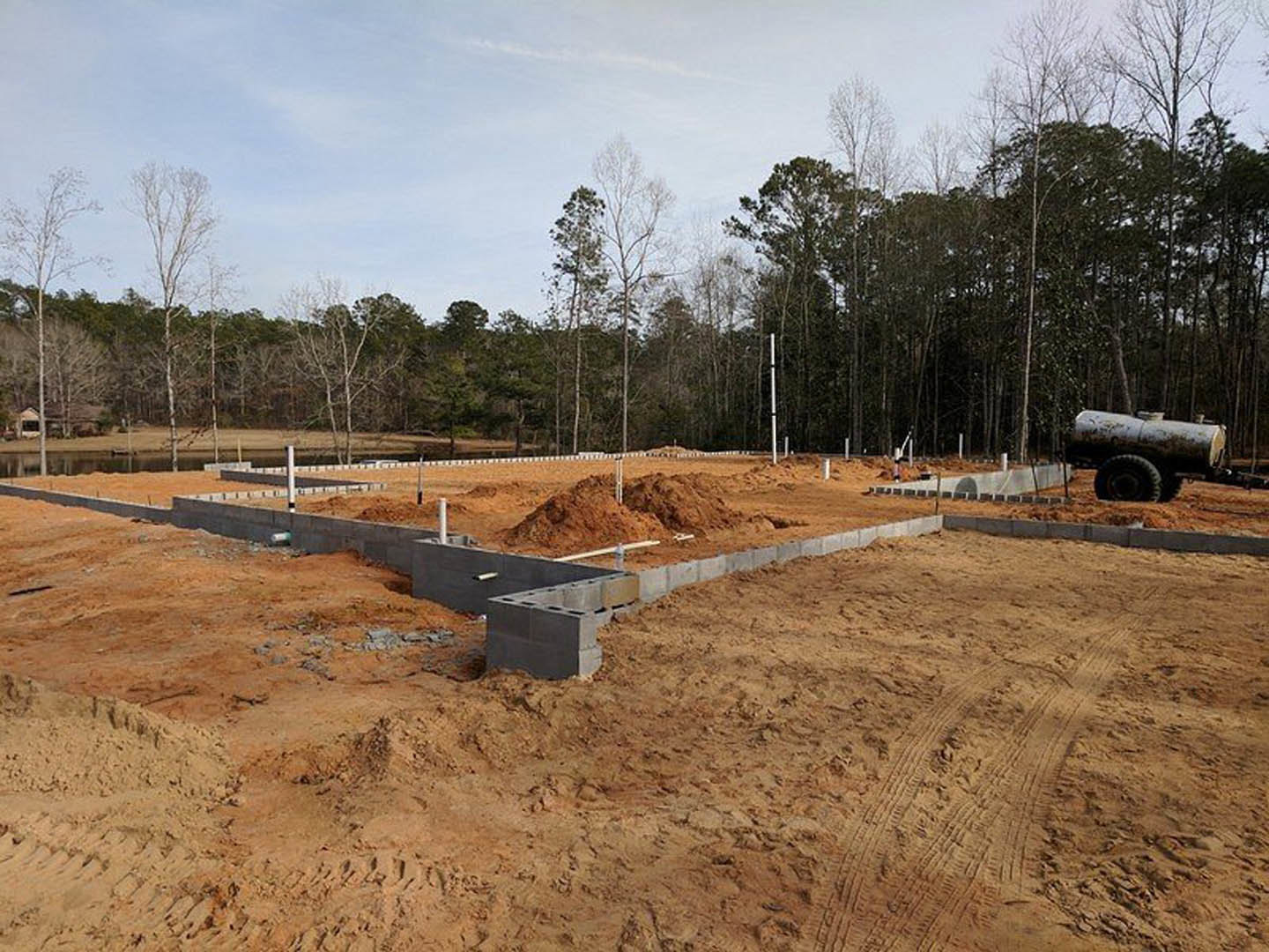 Man standing on dirt construction site in front of partially built home with exposed foundation, concrete blocks, and scattered building materials; trees and grassy field in
