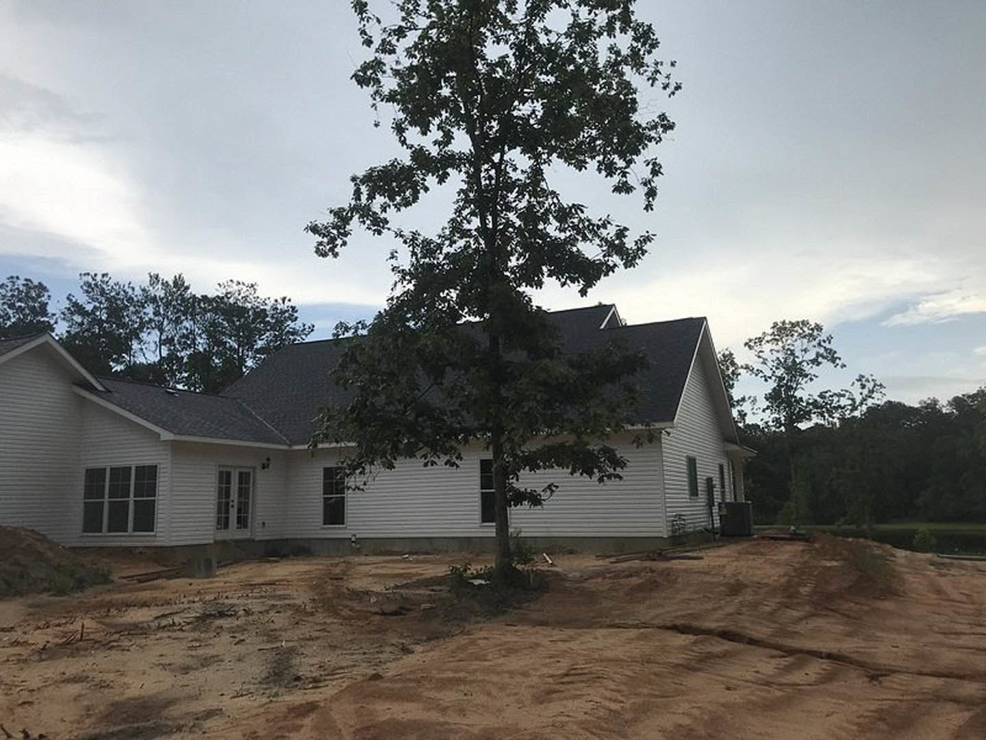 Partially built home with exposed framing and multi-pane windows, leafy tree in front, dirt yard, cloudy sky, and trees in background