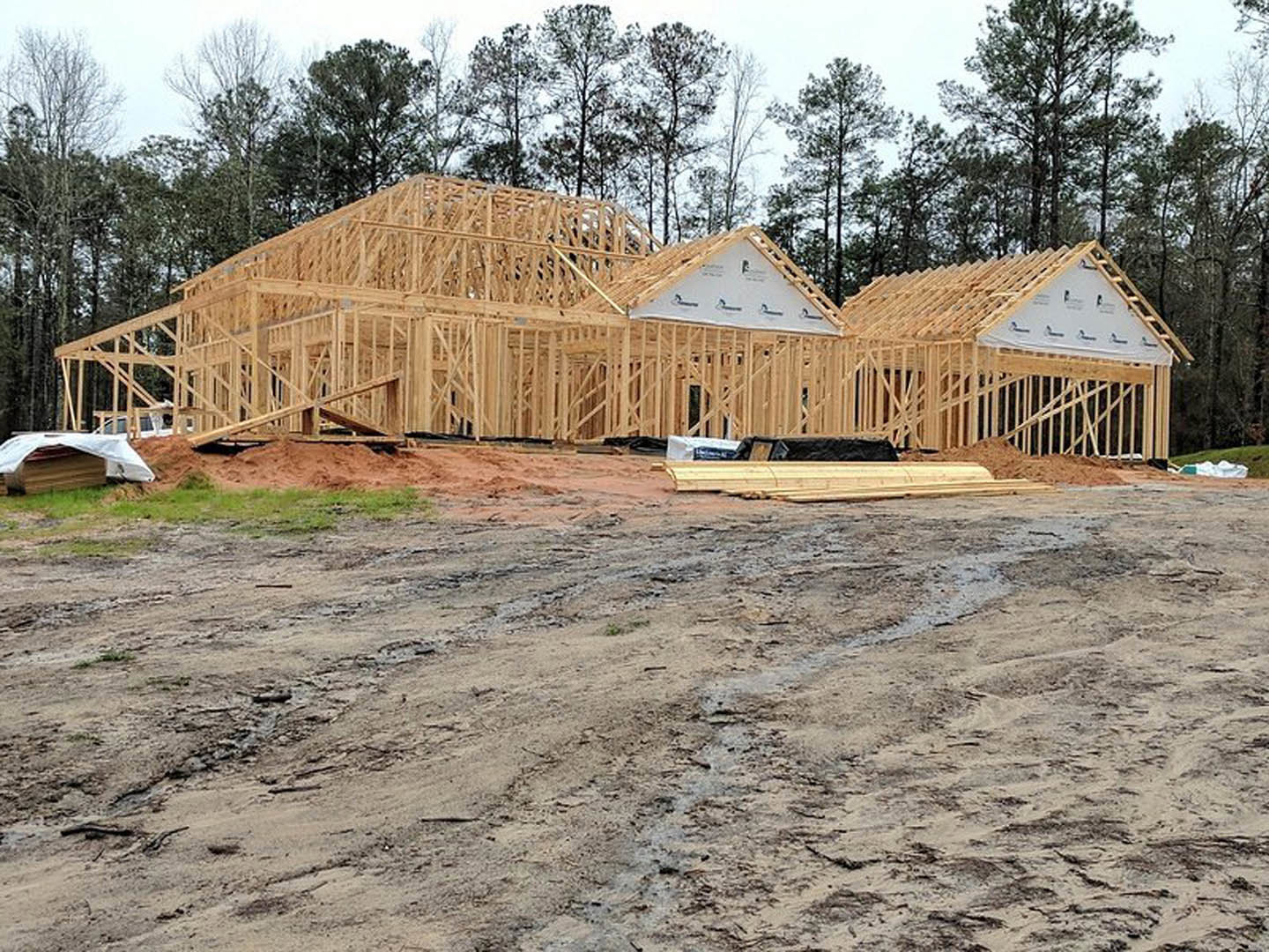 Framed house under construction on a dirt field, surrounded by scattered lumber and trees in the background