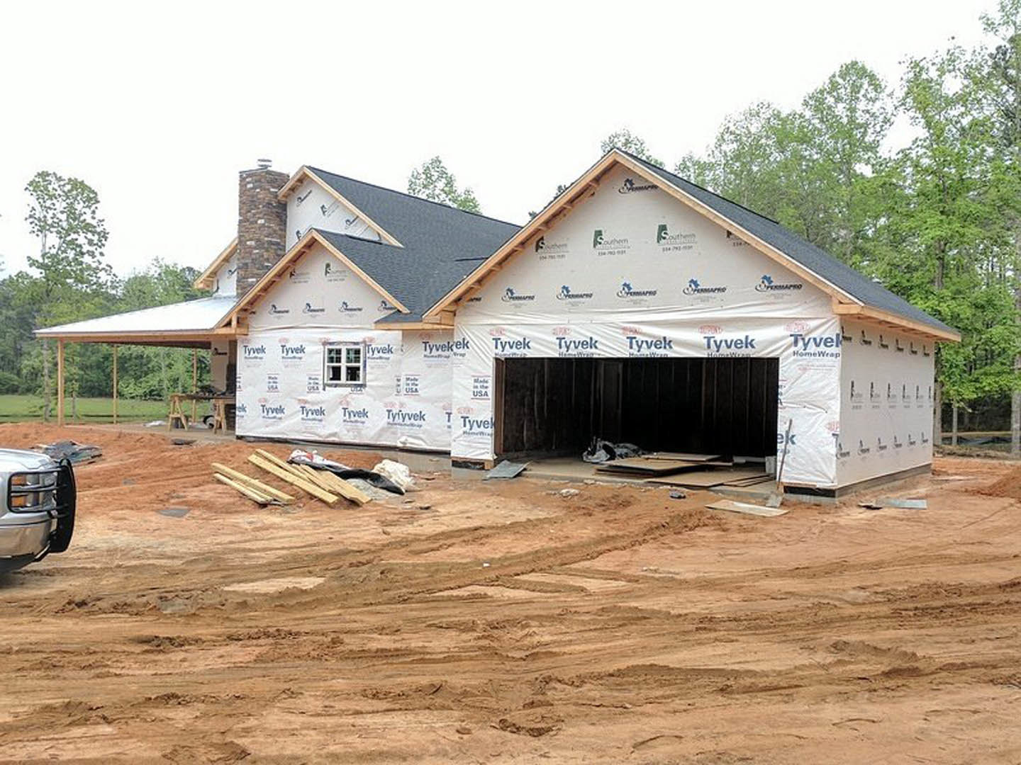 Framed house under construction with attached garage, white sheeting covering exterior walls, pile of lumber on dirt driveway, person lying near front bumper of parked car, trees