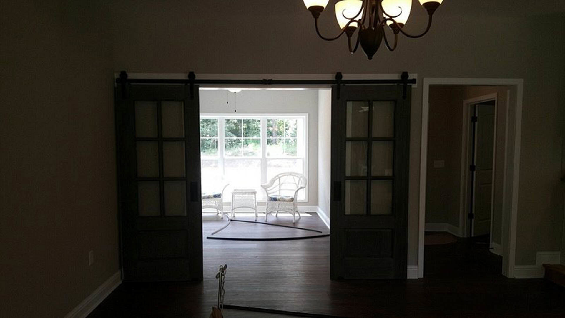 White wicker chair beside sliding glass doors, light wood flooring, modern chandelier overhead, white bench and additional chair in background, black-framed glass door on adjacent