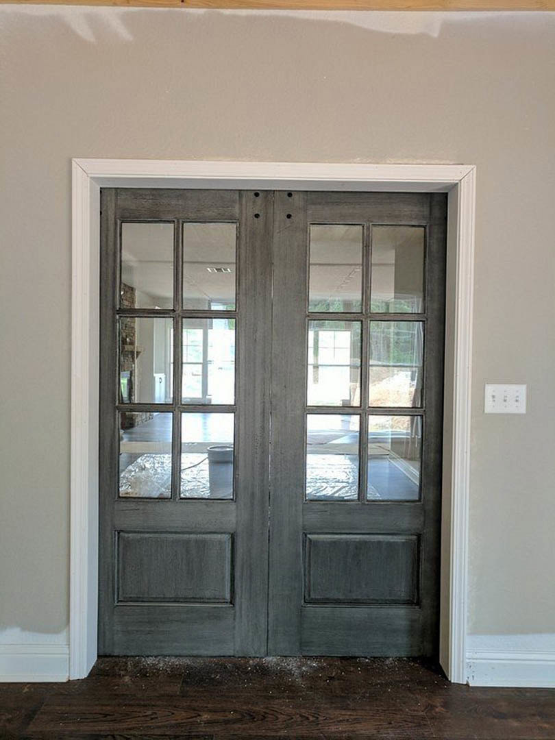 Double glass-paneled double doors with black handles, white wall outlet with black and white buttons, close-up of wood surface, wall with windows, black drawer, and grey