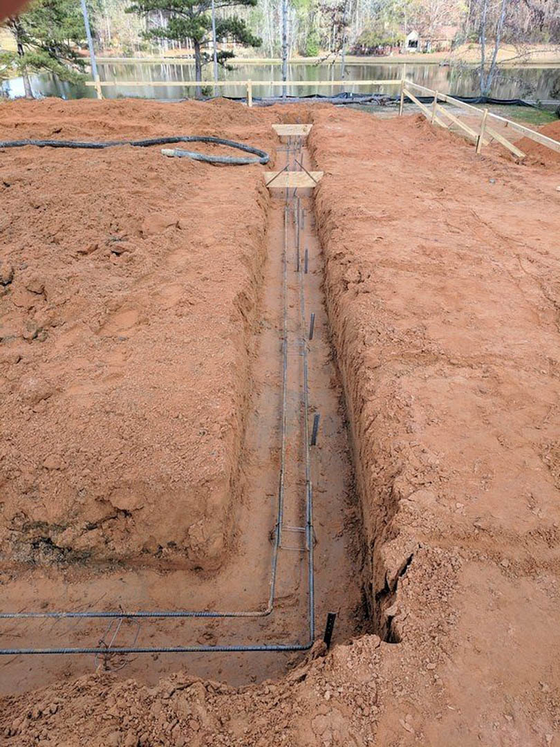 Trench dug in dirt field with exposed pipes, wooden fence in background, large mound of soil with protruding metal rods, scattered plants and trees along perimeter