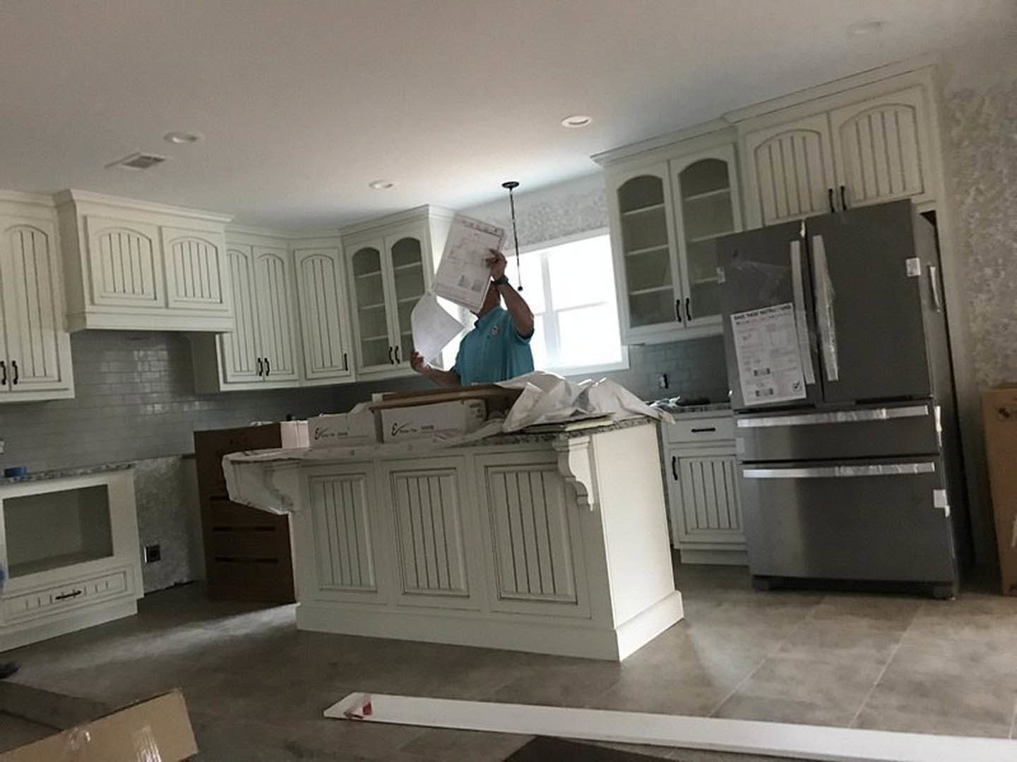 Man holding paper in modern kitchen with white cabinetry, glass-front cabinet, black oven, stainless refrigerator with tape, and light countertops