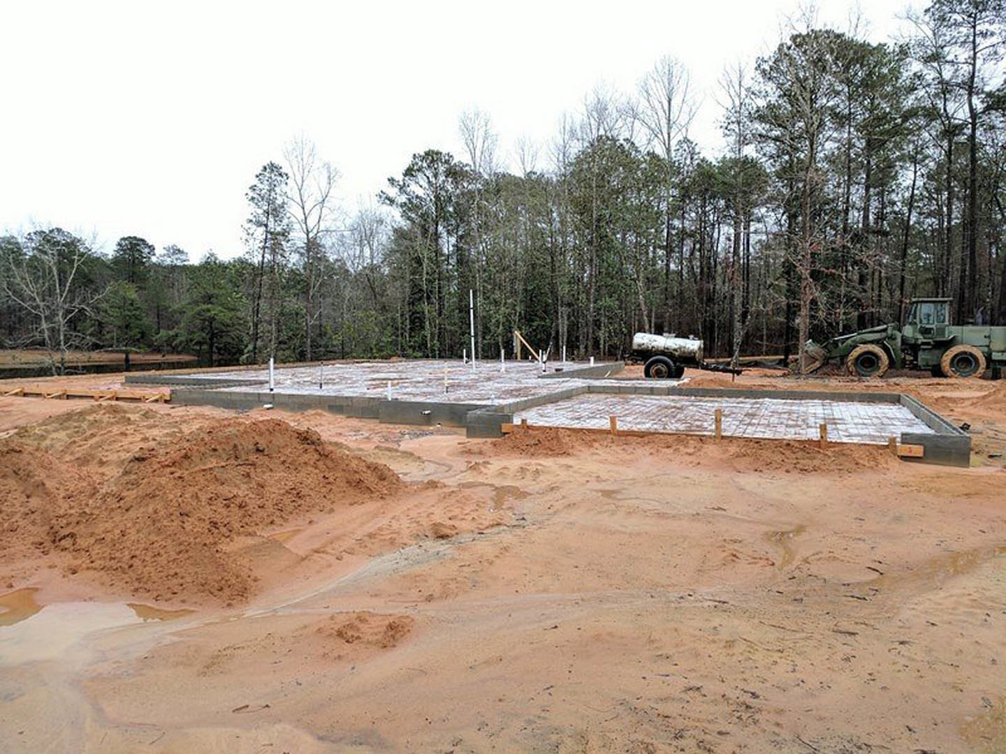 Green tractor with large tires parked on sandy construction site, surrounded by dirt piles, temporary fencing, and dense forest trees in the background