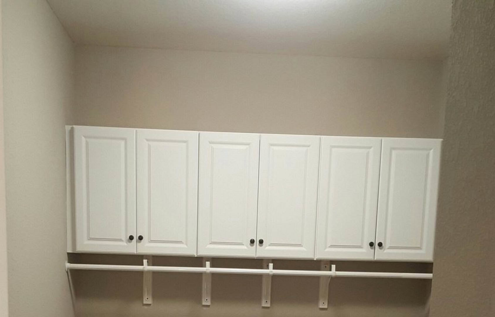 White shaker-style cabinets with black knobs installed along a wall, adjacent to a white door with a black handle and a white metal bar mounted above.