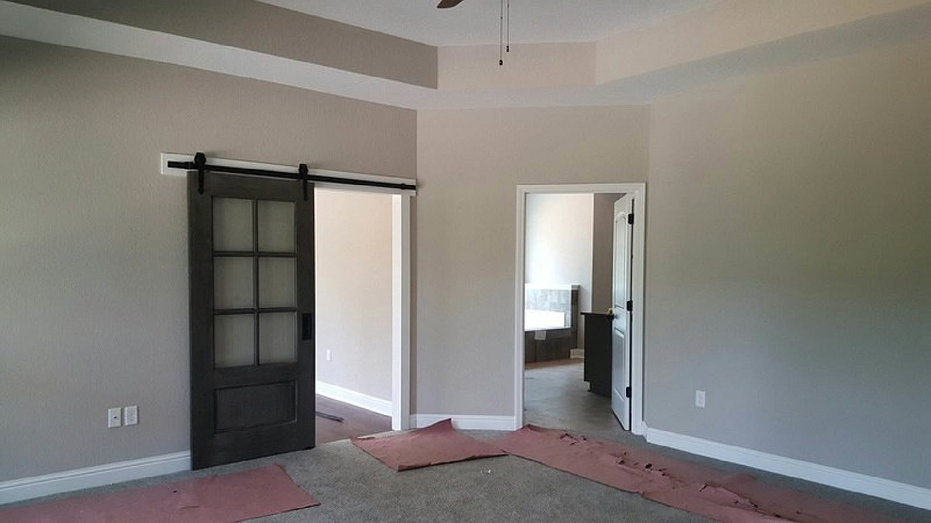 White-walled room with wood plank flooring, ceiling fan, single-panel door, black table, wall outlet, red bag near the floor, and partially visible window