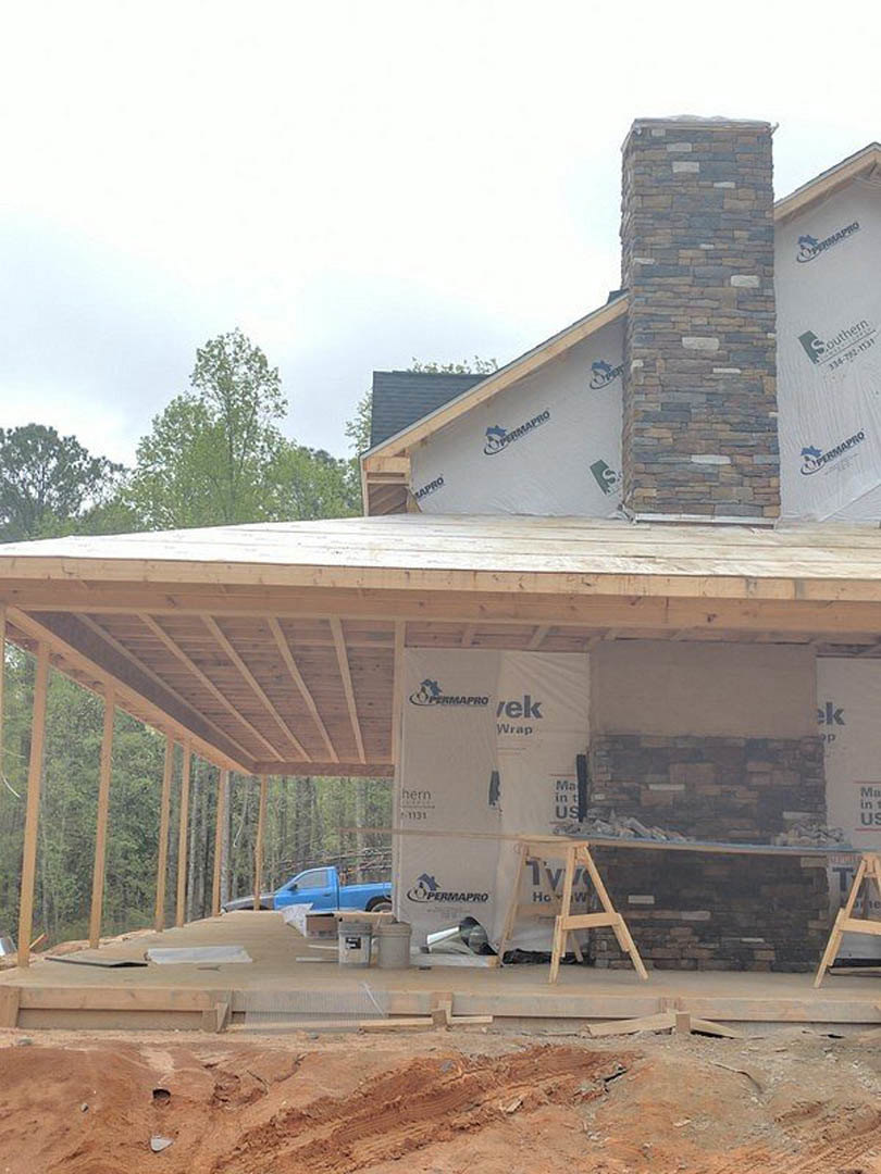 Partially built house with exposed framing, brick chimney, wooden ladder leaning against exterior wall, construction materials scattered on ground, blue truck parked nearby, trees