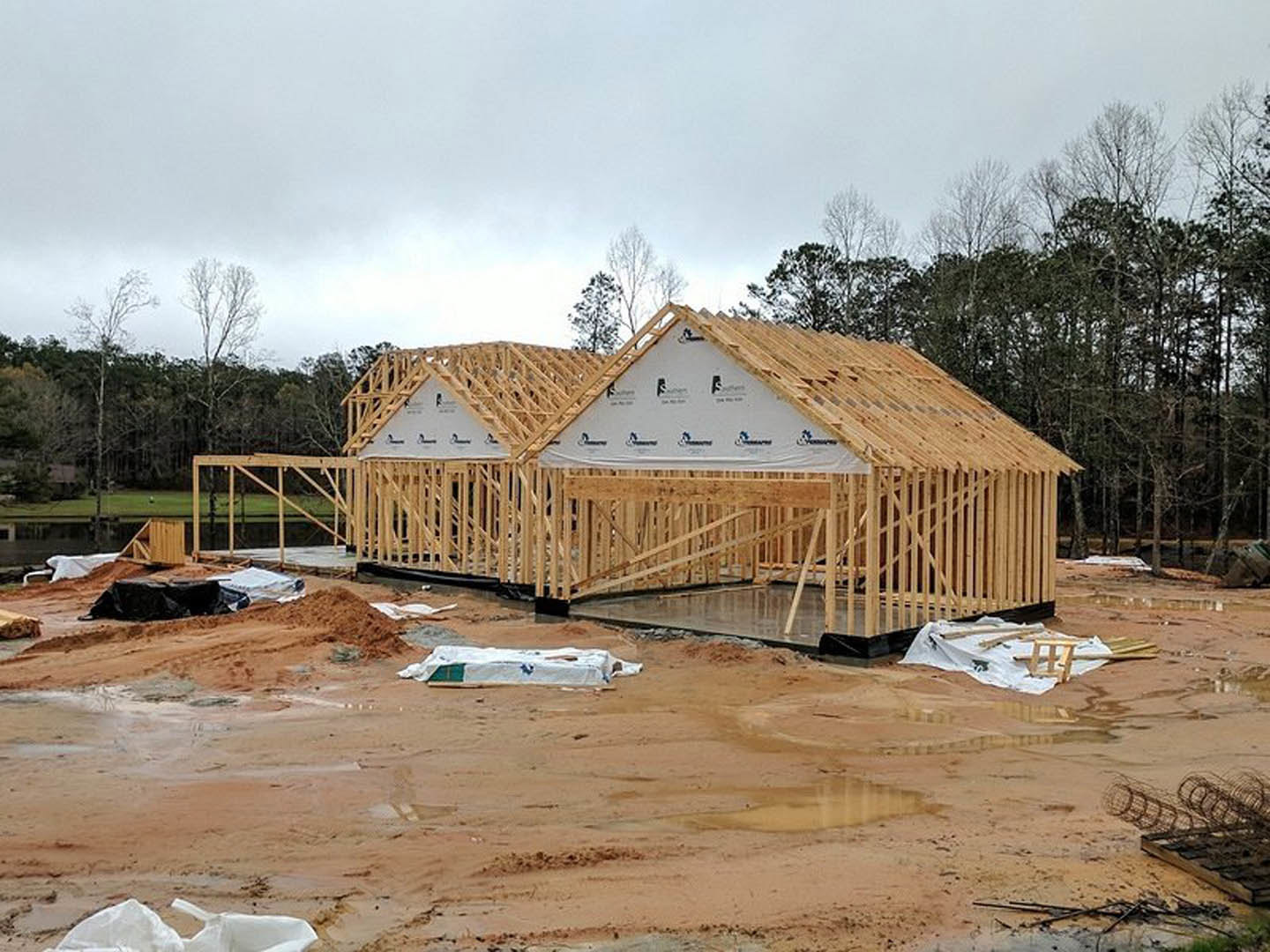 Wood-framed house under construction on dirt lot, surrounded by piles of sand and lumber, partially covered with white sheeting, metal fencing on ground, trees and sky in