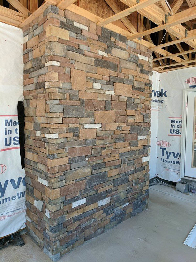 Red and brown brick column under construction, surrounded by unfinished walls and building materials, white paper sign with blue text attached to surface, grey square object nearby