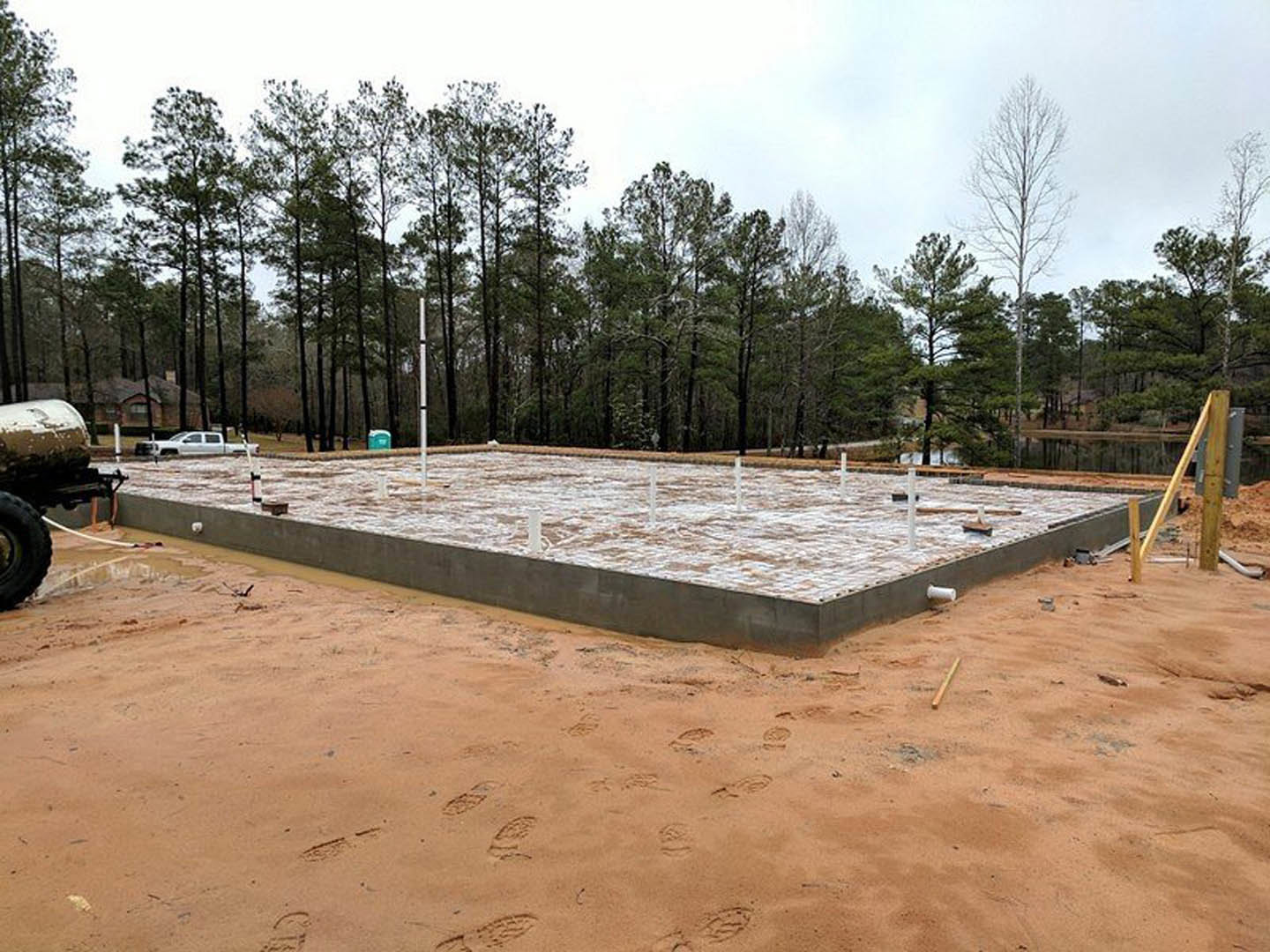 Concrete house foundation surrounded by wet soil, construction materials, wooden posts, and beams, with tire tracks and footprints visible in the sand.